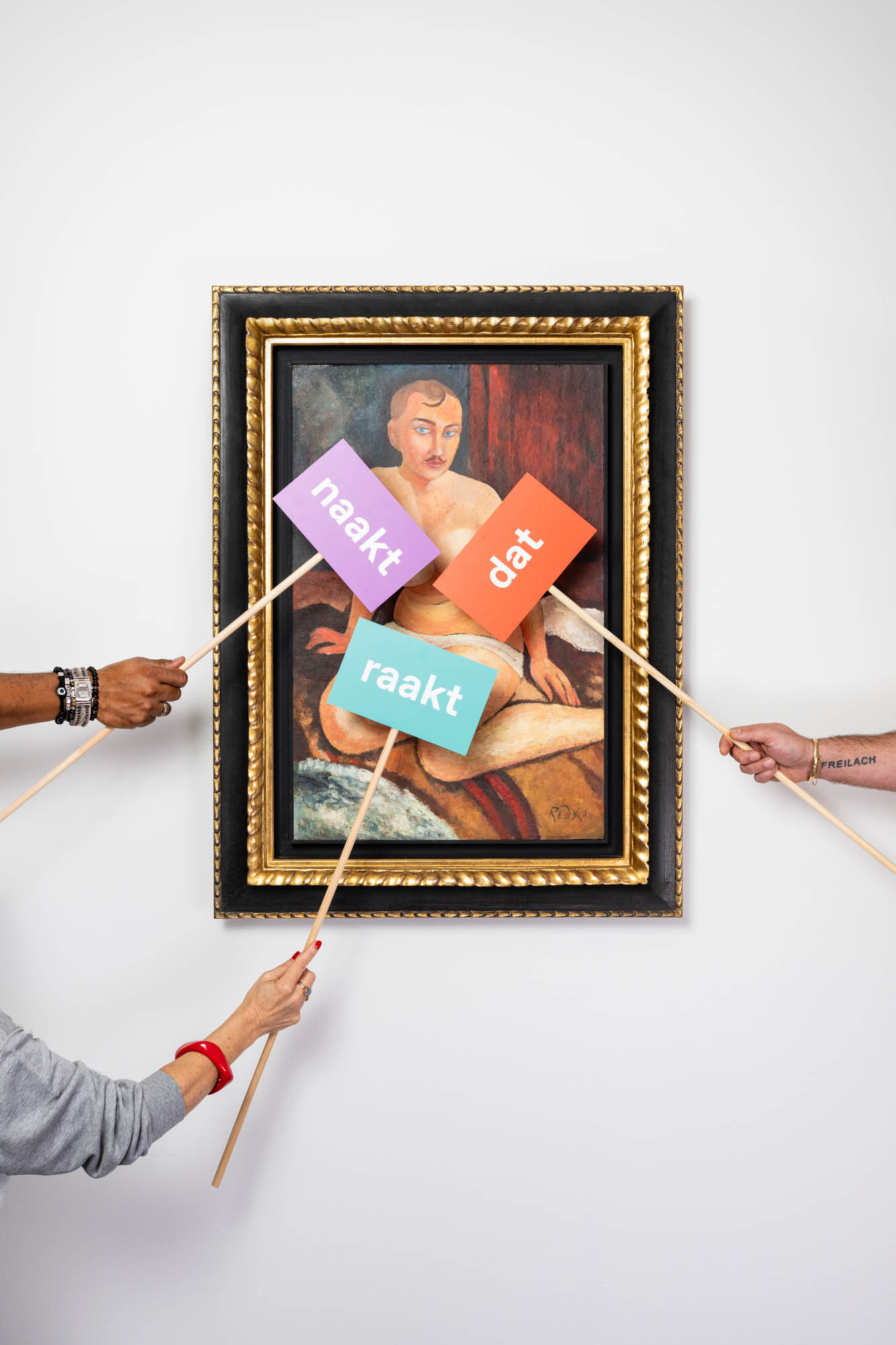 Ingelijst naakt-schilderij waaromheen handen met stokjes de woorden naakt, dat en raakt presenteren